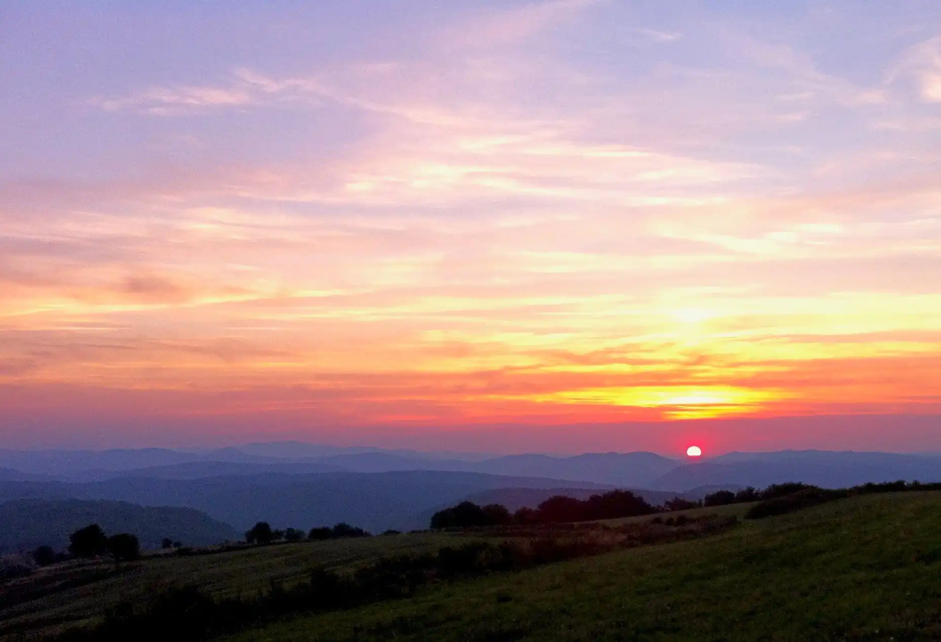 The setting sun casts colorful light across the sky over hilly landscape.